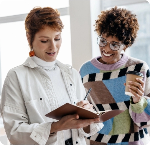 Zwei Frauen stehen nebeneinander und sehen sich Notizen in einem Notizbuch an.