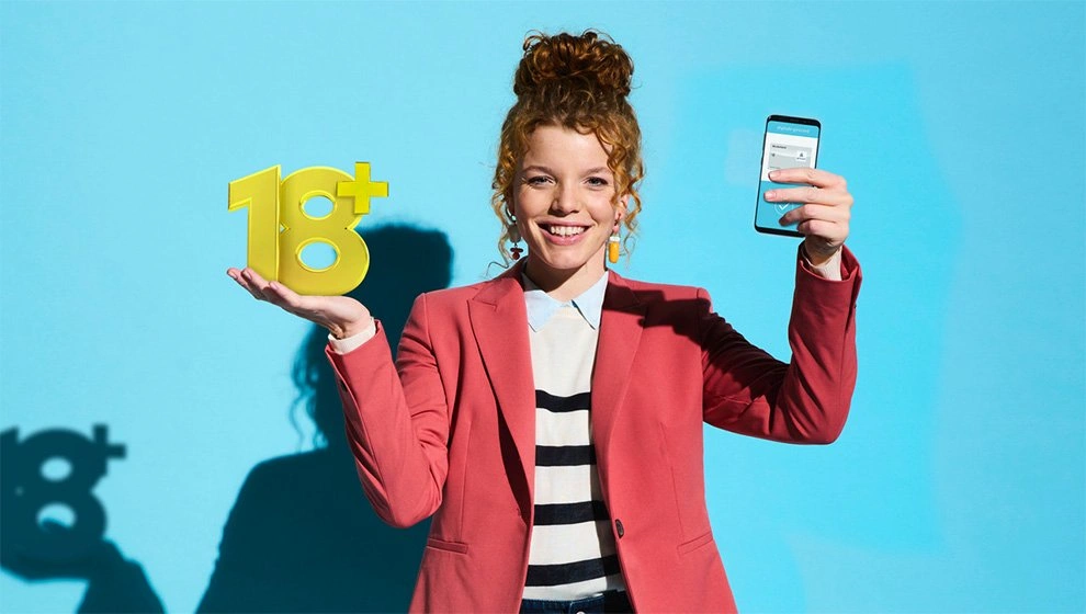 Junge Frau mit lockigem Haar, die ein gelbes 18+ Zeichen in einer Hand und ein Smartphone in der anderen Hand hält, vor einem hellblauen Hintergrund. Sie trägt einen roten Blazer und ein gestreiftes Oberteil und lächelt.