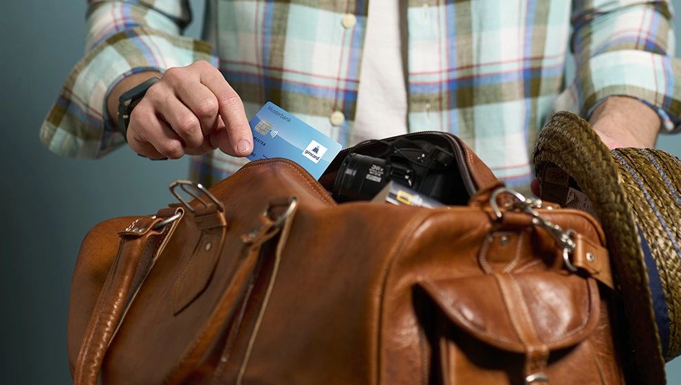Mann mit kariertem Hemd, der eine Kreditkarte aus einer braunen Ledertasche zieht, während er sich auf einen Ausflug vorbereitet. In der Tasche sind auch eine Kamera und ein Strohhut sichtbar.
