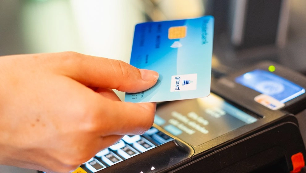 Hand, die eine Kreditkarte an einem Kartenlesegerät hält, während eine Transaktion durchgeführt wird. Die Karte zeigt ein modernes Design und ist für kontaktloses Bezahlen geeignet.