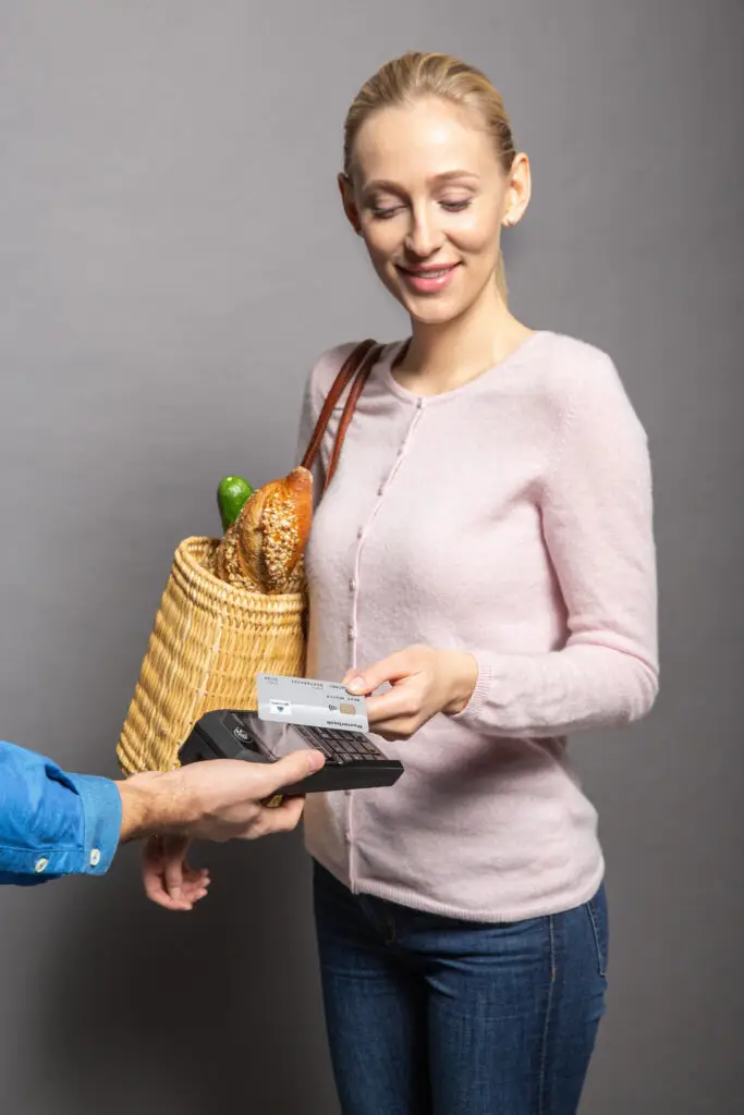 Frau mit einem Korb voller Lebensmittel, die beim Bezahlen mit einer Kreditkarte an einem Kassenterminal lächelt.