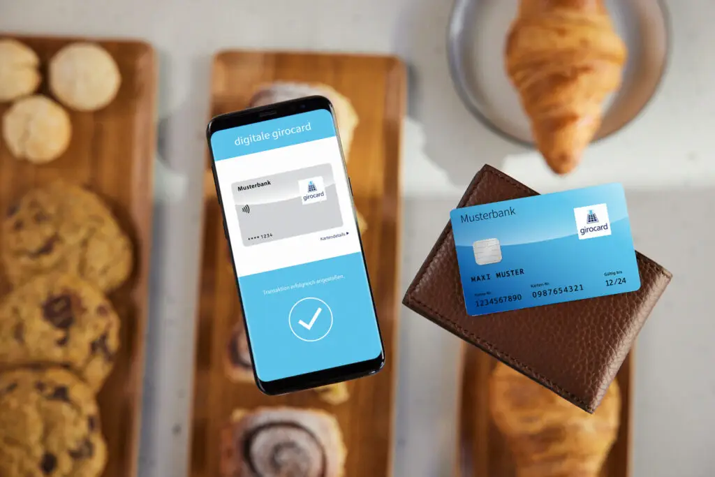 Smartphone mit digitaler Girocard und einer physischen Girocard in einer Geldbörse, umgeben von Gebäck auf einem Holzbrett. Das Bild verdeutlicht die Nutzung moderner Zahlungsmethoden und die Integration von digitalen Finanzlösungen im Alltag.