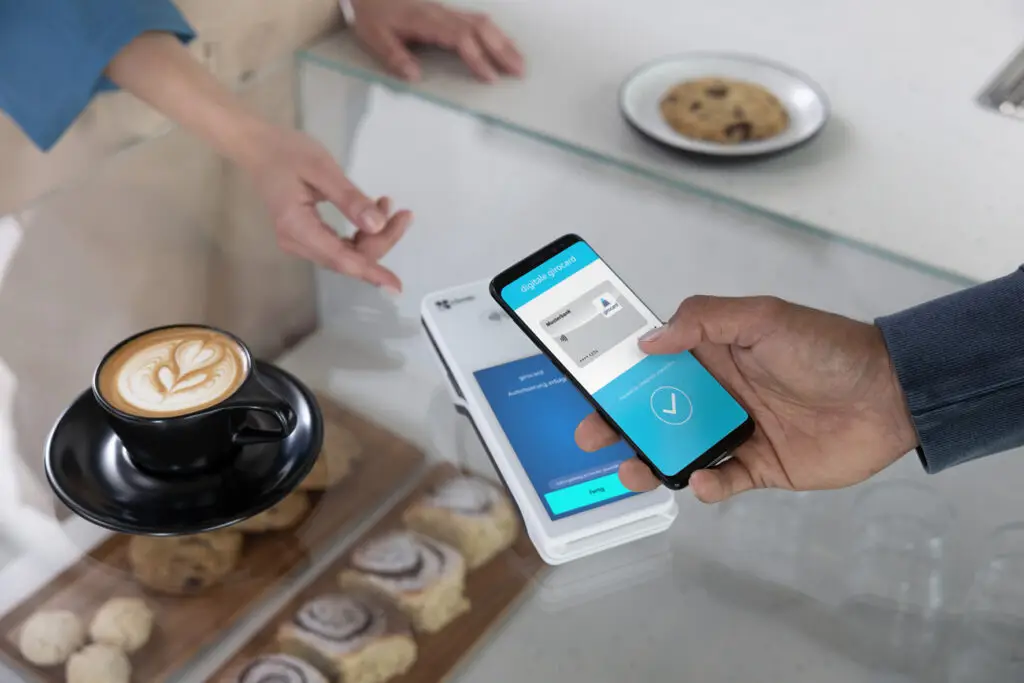 Eine Person nutzt ein Smartphone zur Bezahlung in einem Café, während eine Tasse Kaffee und verschiedene Backwaren auf dem Tisch stehen. Der Bildschirm des Smartphones zeigt eine digitale Gutscheinkarte an, die zur Authentifizierung verwendet wird.