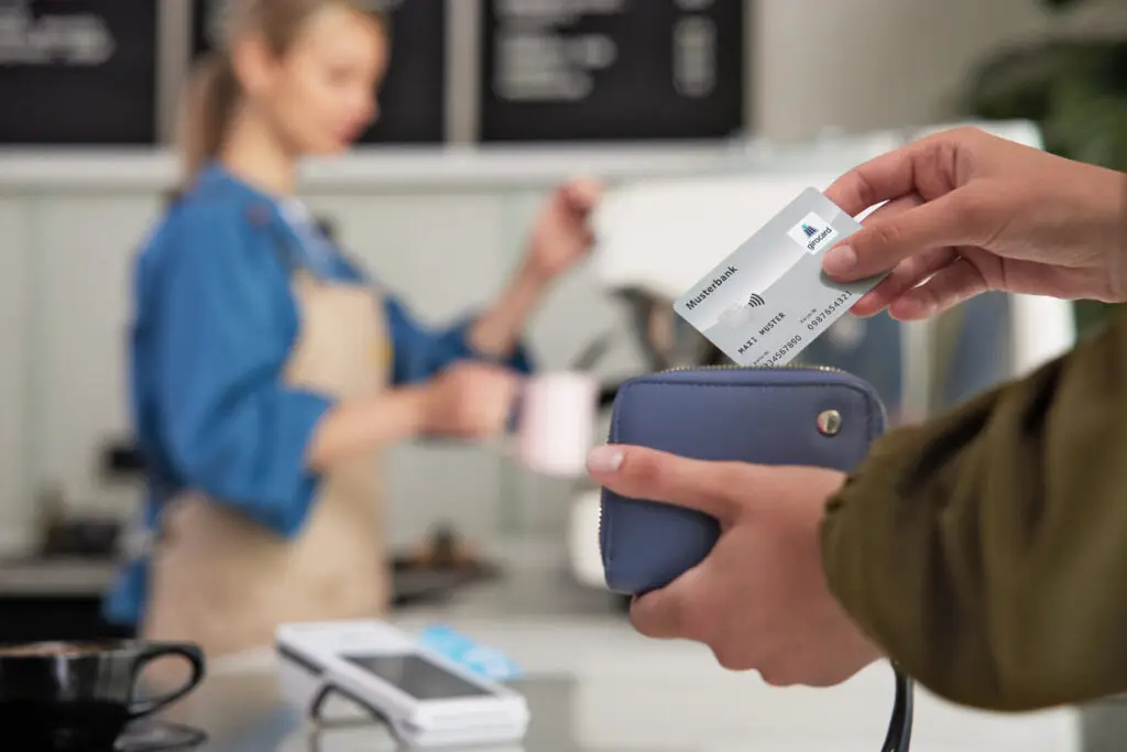 Hand, die eine Kreditkarte aus einer Geldbörse zieht, während im Hintergrund eine Barista einen Kaffee zubereitet. Das Bild zeigt den modernen Zahlungsprozess in einem Café.