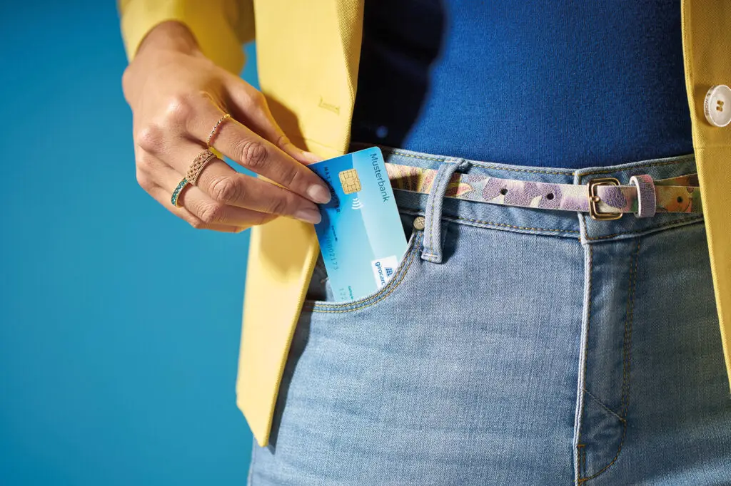 Dame eine Hand, die eine Bankkarte in die Tasche einer Jeans steckt, während sie ein gelbes Blazerjackett trägt, vor einem blauen Hintergrund.