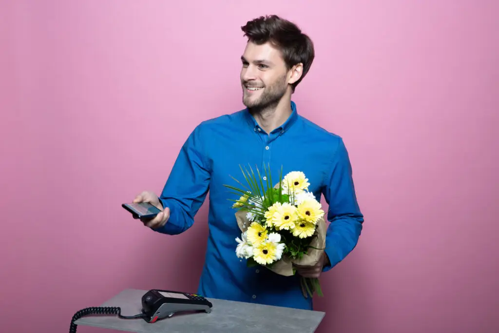 Mann in blauer Hemd hält einen Blumenstrauß mit gelben und weißen Blumen in einer Hand und ein Smartphone in der anderen, während er an einem Tisch mit einem Kassengerät steht, vor einem pinken Hintergrund.