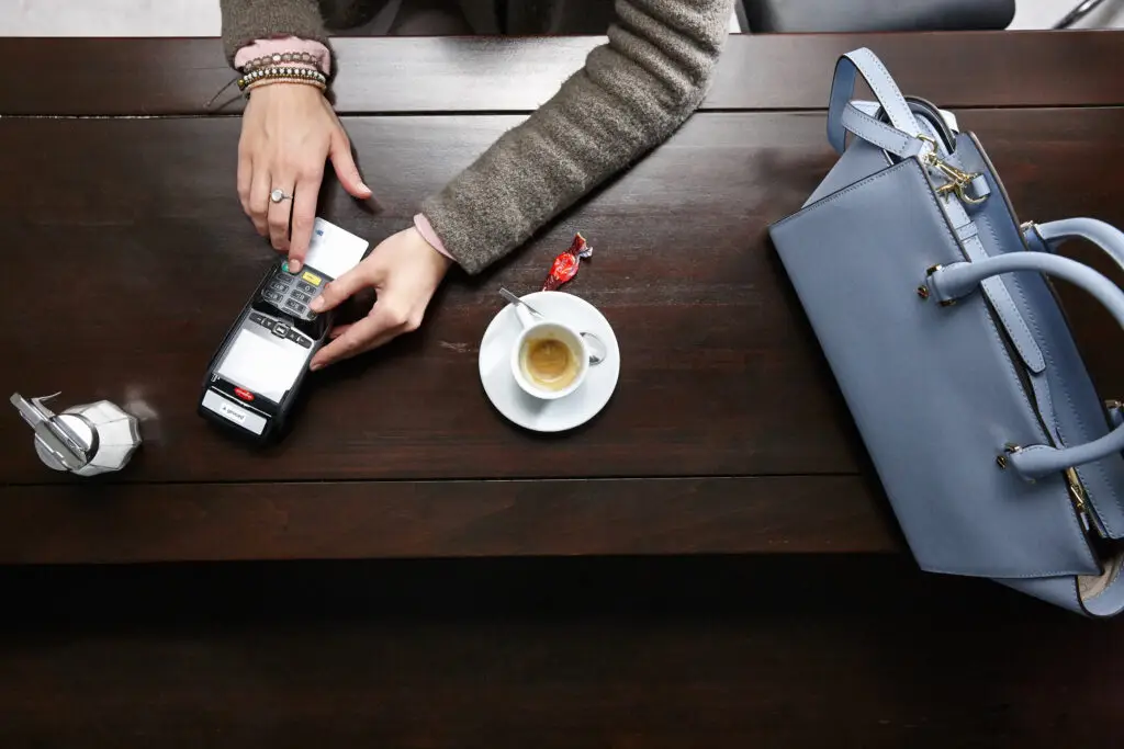 Eine Person bezahlt mit einer Kreditkarte an einem modernen Kartenlesegerät, während eine Tasse Espresso auf einem Tisch steht. Neben der Tasse liegt ein Löffel und ein Stückchen Süßigkeit. Eine stilvolle blaue Handtasche ist ebenfalls auf dem Tisch zu sehen.