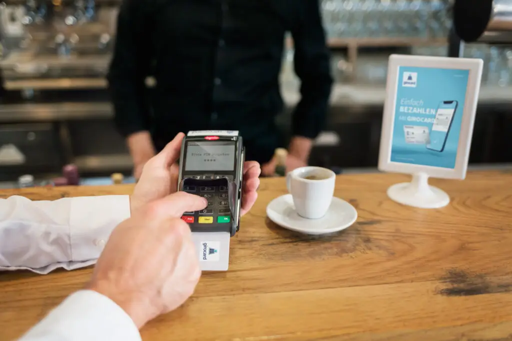 Kunde tätigt eine Zahlung mit einem Kartenlesegerät in einem Café, während eine Tasse Kaffee auf dem Tisch steht und ein Schild mit Informationen zum Bezahlen mit Girocard sichtbar ist.