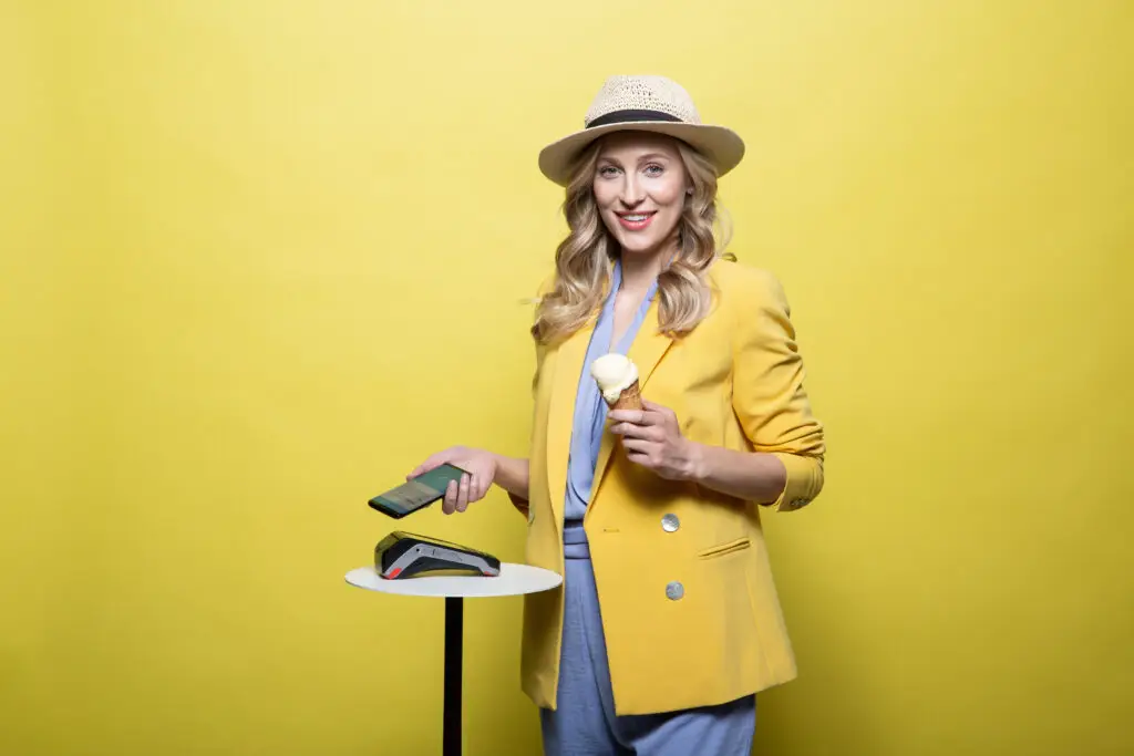 Frau in gelbem Blazer und Strohhut hält ein Eis und ein Smartphone, steht neben einem Zahlungsterminal vor einem gelben Hintergrund.