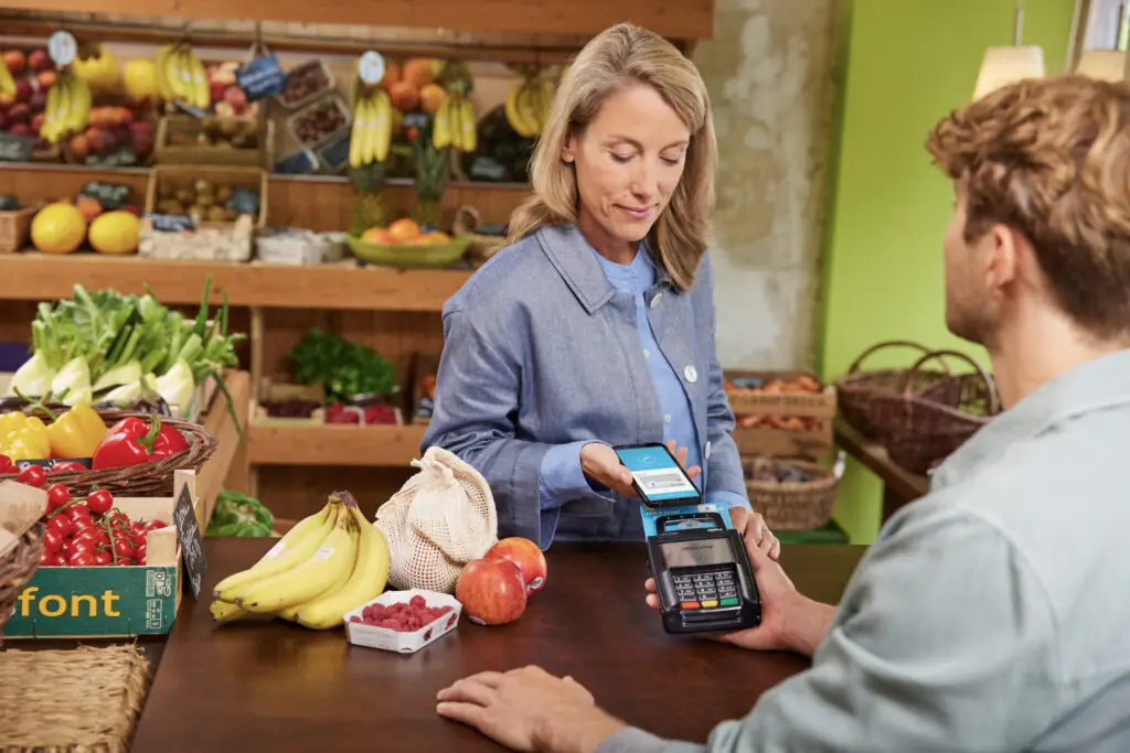 Eine Frau scannt mit ihrem Smartphone eine Zahlung an einem Kassenterminal in einem Lebensmittelgeschäft, während frisches Obst und Gemüse im Hintergrund sichtbar sind.