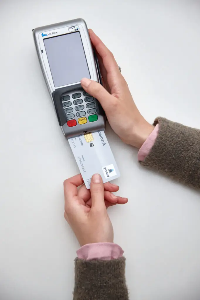 Hände, die eine Kreditkarte in ein Kartenlesegerät einführen, um eine kontaktlose Zahlung durchzuführen. Das Gerät zeigt eine leere Anzeige und befindet sich auf einem hellen Tisch.