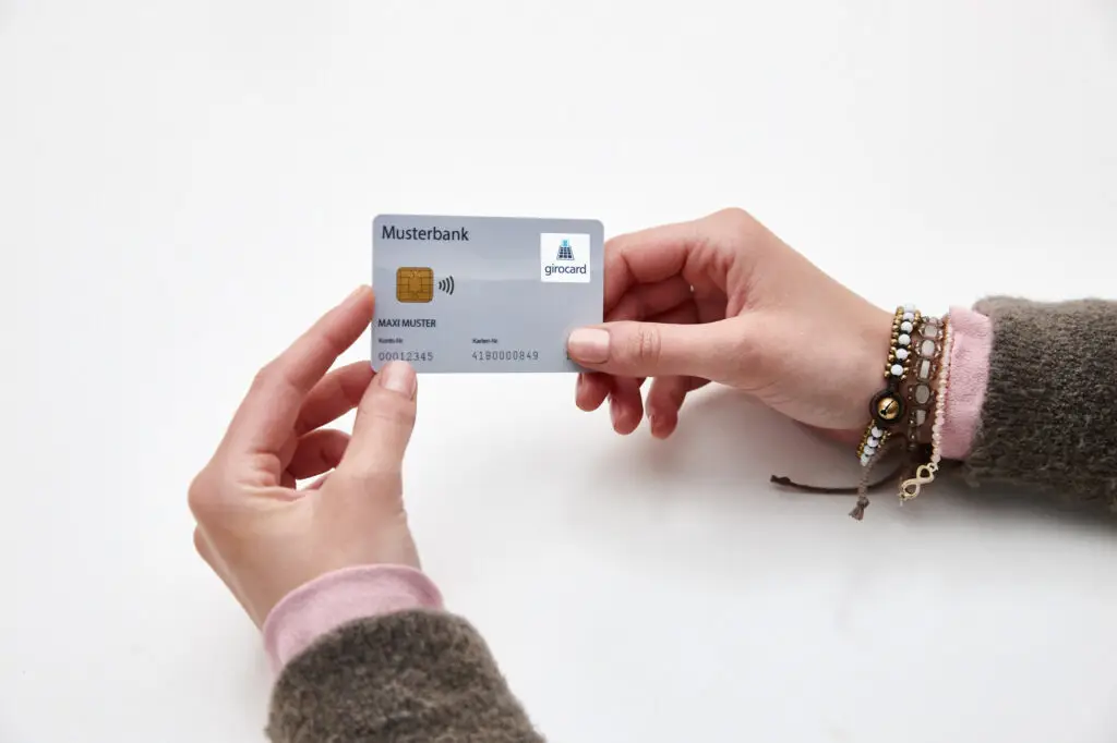 Eine Person hält eine girocard der Musterbank in der Hand, die mit einem Chip und einem kontaktlosen Zahlungssymbol ausgestattet ist. Die Karte zeigt den Namen des Kontoinhabers und die Kontonummer. Im Hintergrund sind zarte Armbänder sichtbar, die den persönlichen Stil unterstreichen.