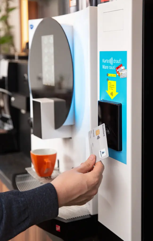 Nutzer scannt eine Karte an einem modernen Verkaufsautomaten, um eine Ware zu entnehmen. Im Hintergrund ist eine orangefarbene Tasse zu sehen, die auf der Ablage steht. Der Automat zeigt eine Anleitung zur Kartennutzung.