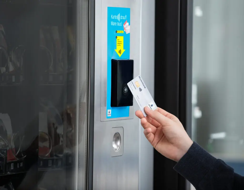 Hand, die eine Karte an ein kontaktloses Zahlungsterminal eines Automaten hält, mit der Aufforderung, die Karte aufzulegen, um eine Ware zu entnehmen. Im Hintergrund sind verschiedene Produkte im Automaten sichtbar.