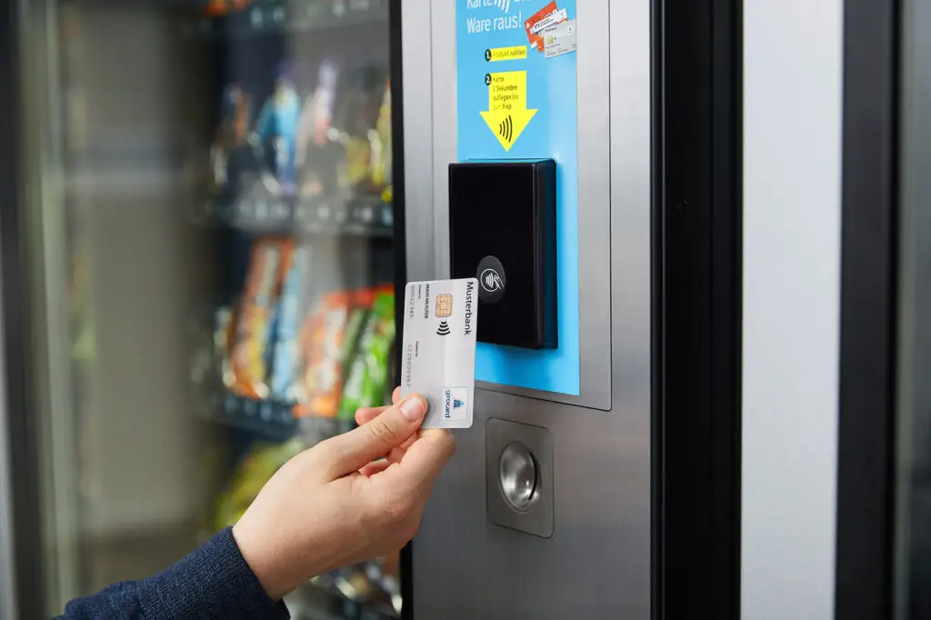 Eine Person hält eine kontaktlose Bankkarte vor einen modernen Snackautomaten, der eine Auswahl an Snacks im Hintergrund zeigt. Der Automat ist mit Anweisungen zur Kartennutzung versehen, um den Kaufprozess zu erleichtern.