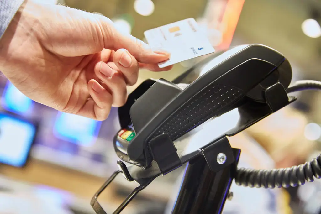 Hand hält eine Kreditkarte, während sie an einem Kartenzahlungsgerät an der Kasse gescannt wird, um eine bargeldlose Transaktion durchzuführen.