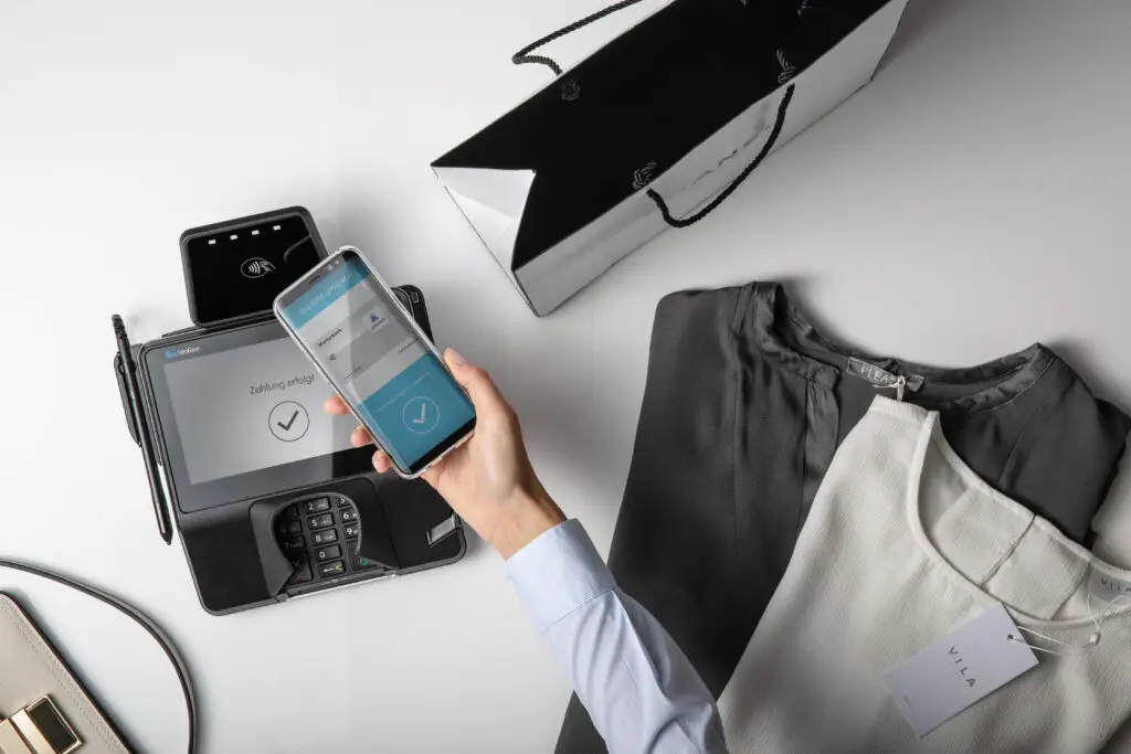 Hand mit Smartphone, das eine kontaktlose Zahlung an einem Kartenlesegerät zeigt, neben einer Einkaufstasche und zwei Oberteilen. Ideal für Online-Shopping und moderne Zahlungsmethoden.