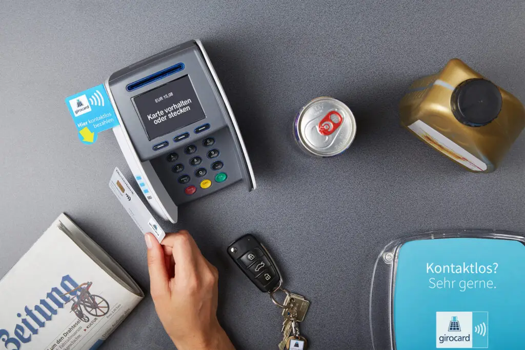 Kassenterminal mit kontaktloser Zahlungsmöglichkeit, einer Hand, die eine girocard hält, einer Getränkedose und einem Schlüsselbund auf einem Tisch.