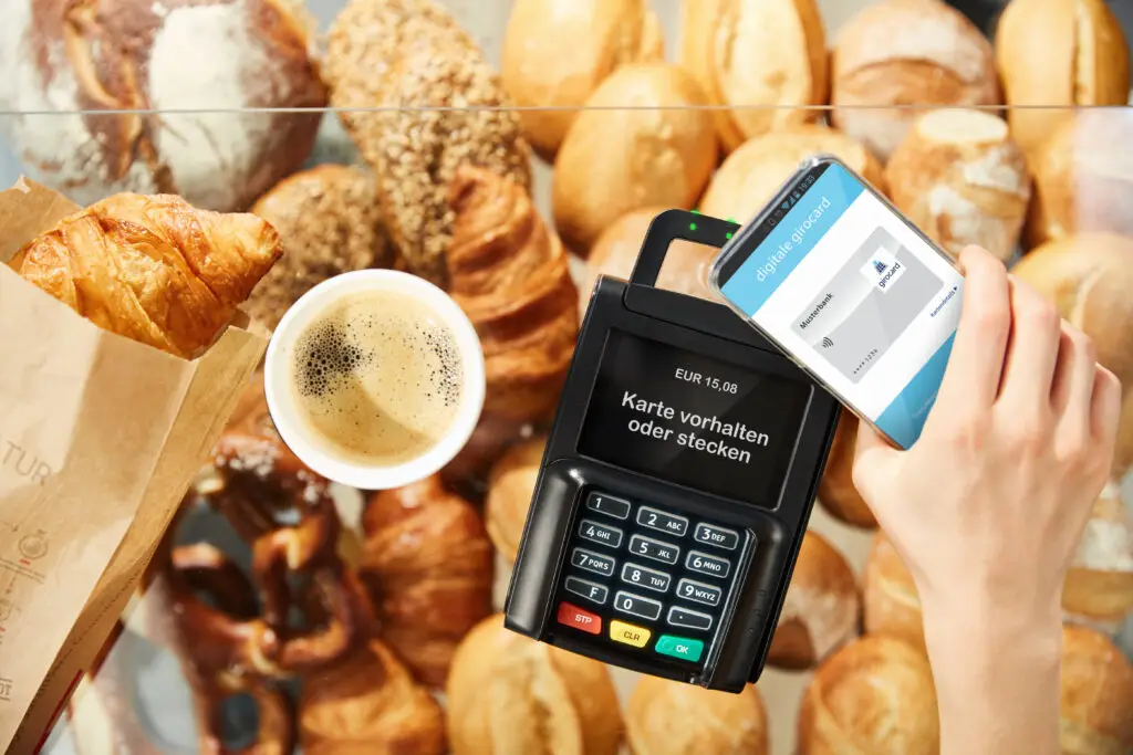 Zahlungsvorgang mit einem Kartenlesegerät und einem Smartphone zur kontaktlosen Bezahlung, umgeben von frischen Backwaren und einer Tasse Kaffee.