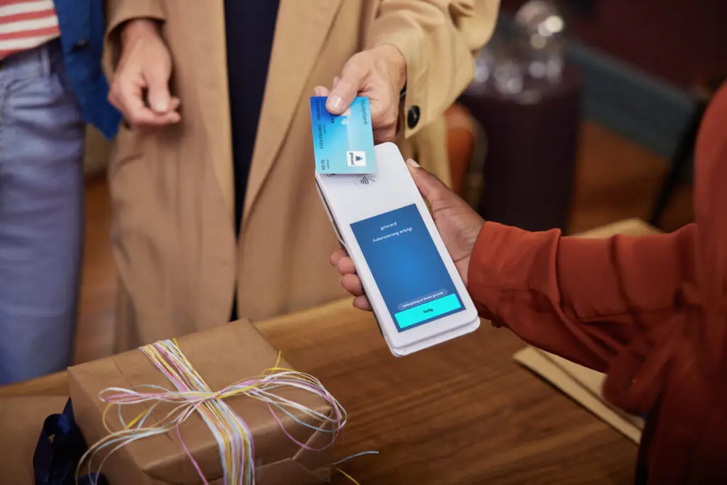 Zahlungsvorgang mit Kreditkarte an einem mobilen Zahlungsterminal, während eine Person ein Geschenk auf einem Tisch hält. Der Bildschirm zeigt eine Bestätigung der erfolgreichen Autorisierung.