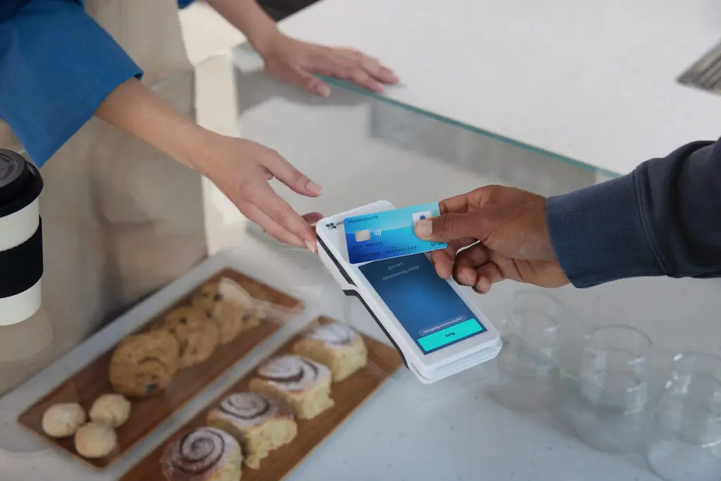 Zahlung mit Karte an einem modernen Kassensystem in einem Café, während eine Person Gebäck auf einem Holzbrett serviert.