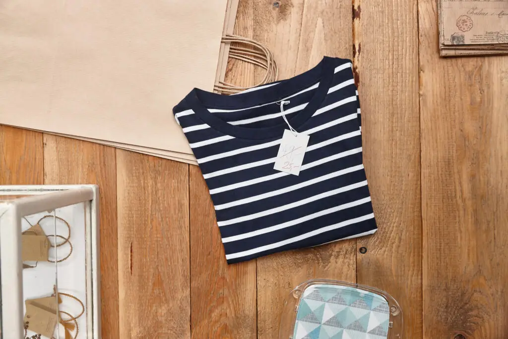 Gestreiftes T-Shirt in Navy-Blau mit weißen Streifen auf einem Holztisch, umgeben von Dekorationsmaterialien und einem Preisetikett von 25 Euro.