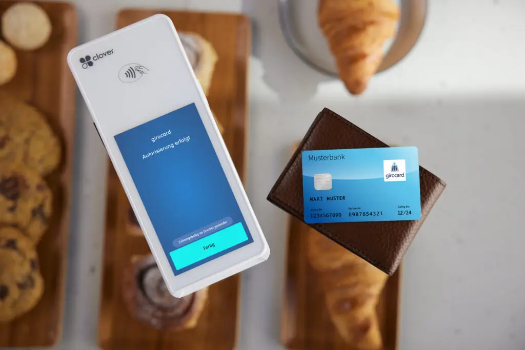 Mobile Zahlung mit girocard auf einem Clover-Terminal, das eine erfolgreiche Autorisierung anzeigt, während eine Bankkarte aus einem Geldbeutel gehalten wird, im Hintergrund Gebäck auf einem Holzbrett.