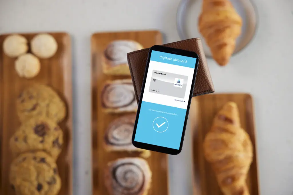 Smartphone mit digitaler Girocard-Anzeige in einer Brieftasche, umgeben von frisch gebackenem Gebäck auf Holzplatten, ideal für die Nutzung moderner Zahlungsmethoden in Cafés oder Bäckereien.