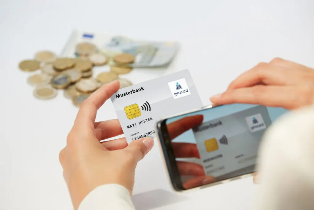 Hand, die eine Girokarte einer Musterbank hält, während ein Smartphone die Karte scannt. Im Hintergrund sind Münzen und Geldscheine sichtbar, was auf finanzielle Transaktionen hinweist.