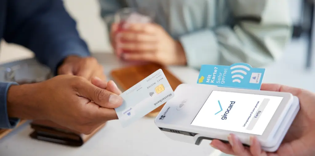 Zwei Personen führen eine kontaktlose Zahlung durch, während eine Hand eine girocard an ein Zahlungsterminal hält. Im Hintergrund sind ein Glas und eine Geldbörse sichtbar, was eine entspannte Cafésituation vermittelt.