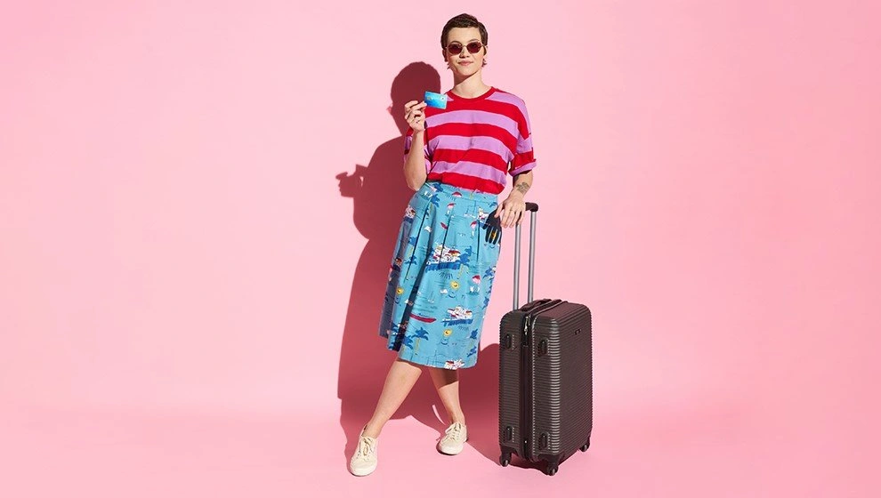 Frau mit Sonnenbrille trägt gestreiftes T-Shirt und einen bunten Rock, steht neben einem Koffer vor einem pinken Hintergrund und hält ein Getränk in der Hand. Ideal für Reise- und Modeinspiration.