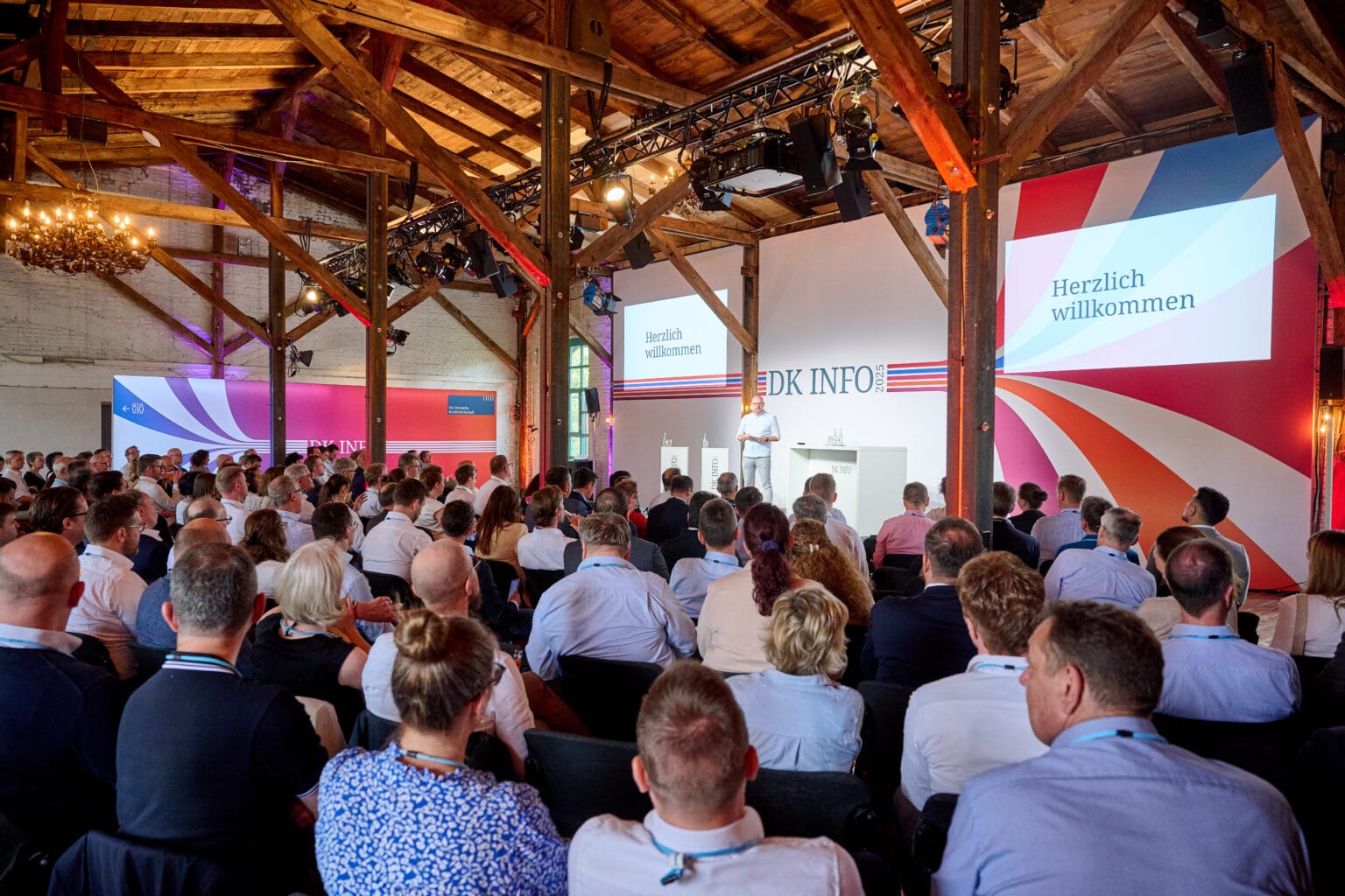 Konferenz im Jahr 2025 mit dem Titel DK INFO, bei der ein Redner auf der Bühne eine Präsentation hält. Im Hintergrund begrüßt ein großes Display die Teilnehmer mit den Worten "Herzlich willkommen". Der Raum ist voll mit Zuhörern, die auf das Geschehen auf der Bühne fokussiert sind.