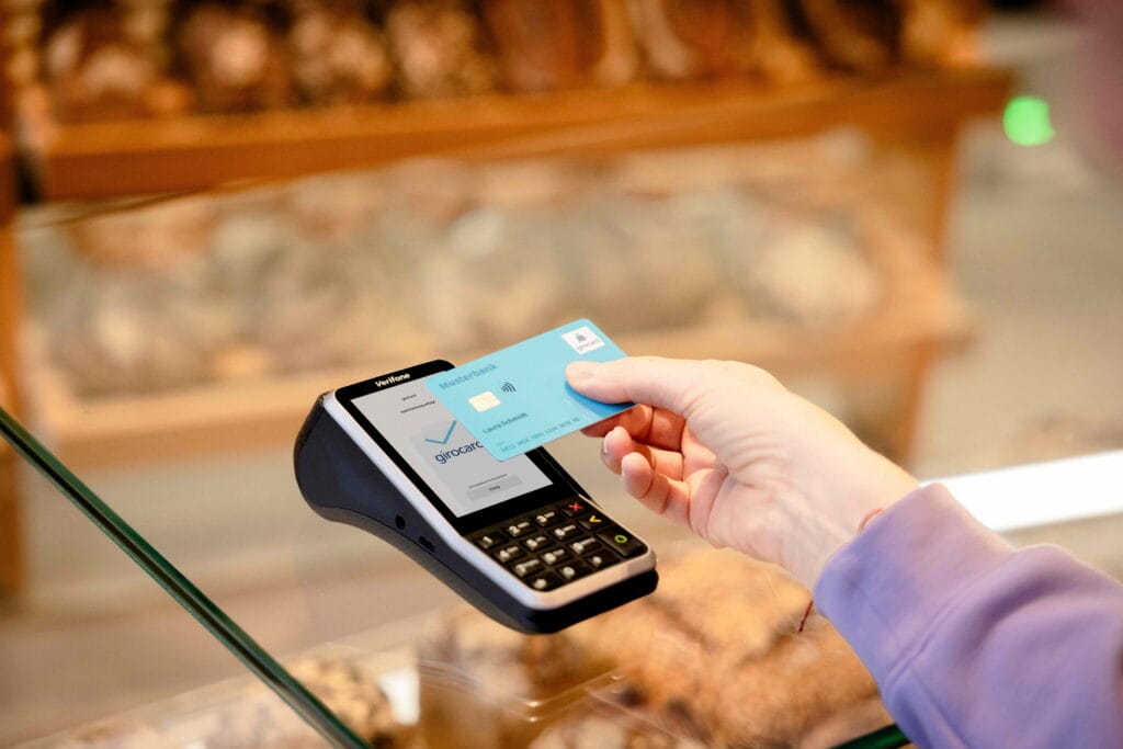 Girocard-Bezahlvorgang: Eine Hand hält eine blaue girocard an ein mobiles Kartenterminal; Hintergrund wirkt wie eine Laden- oder Bankfiliale in warmer Beleuchtung.