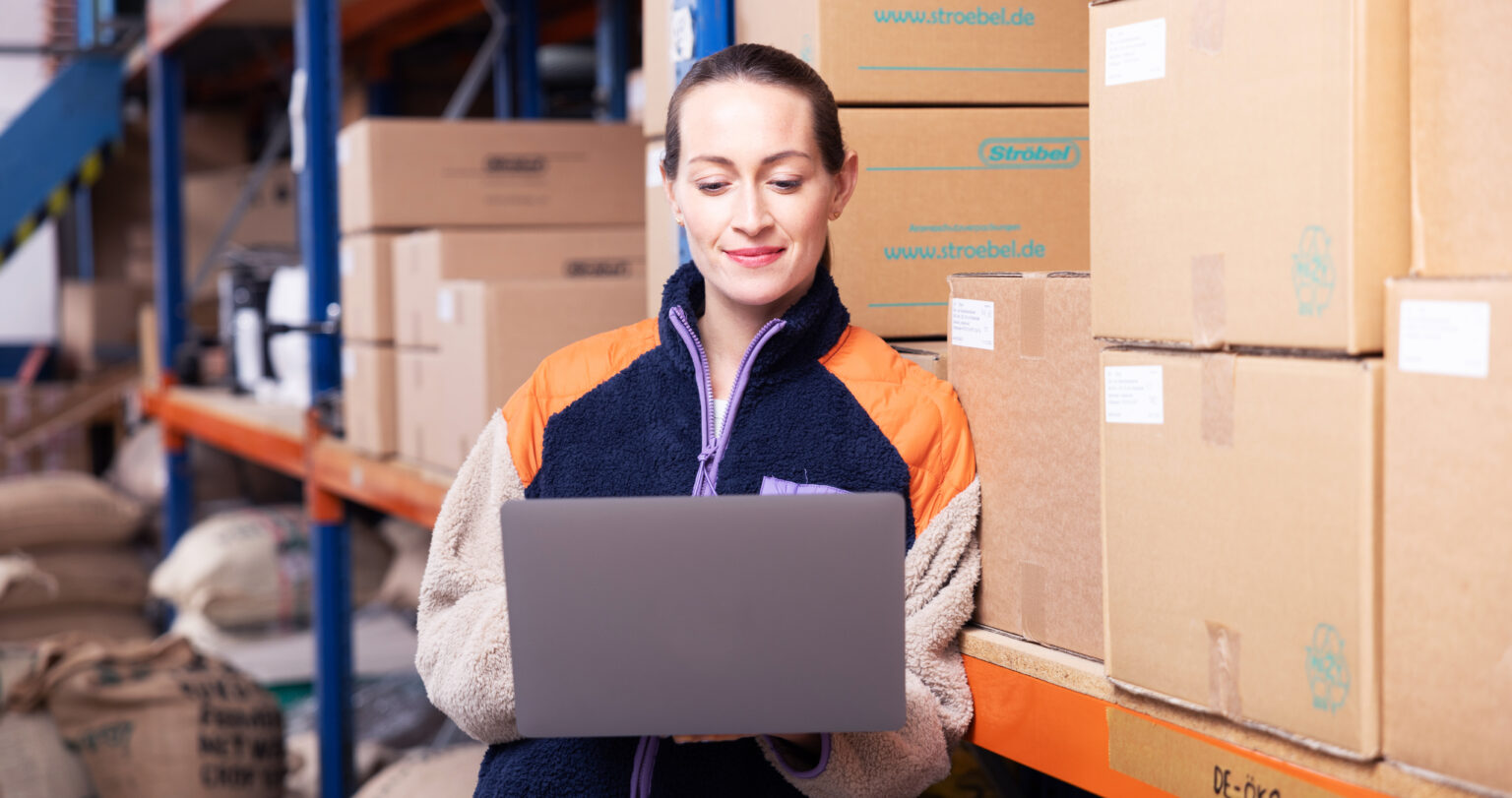 Frau mit Laptop in einem Lager, umgeben von Kartons und Säcken, während sie effizient arbeitet und Informationen verwaltet.
