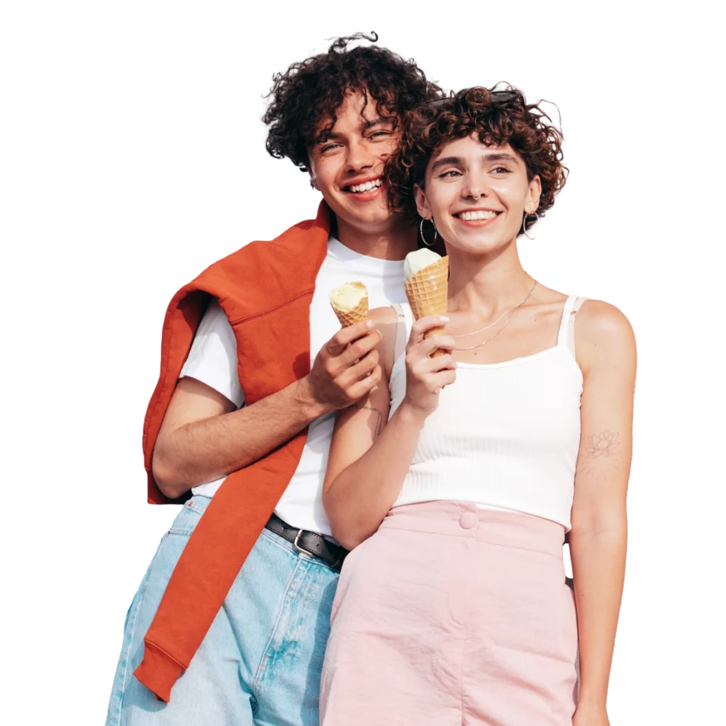 Junges Paar mit lockigem Haar, das fröhlich mit Eiscreme-Waffeln in der Hand posiert, im Freien bei sonnigem Wetter. Die Frau trägt ein weißes Oberteil und einen rosa Rock, der Mann einen roten Pullover über der Schulter und eine lässige Jeans.
