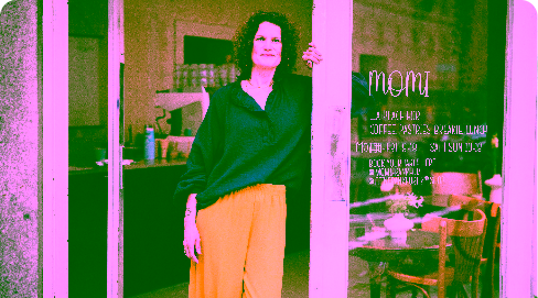 Eine Unternehmerin mit blauer Bluse und gelber Hose lehnt am Türrahmen ihres Geschäfts.