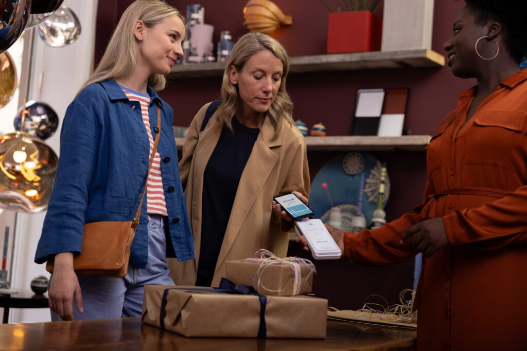 Zwei Frauen stehen in einem modernen Geschäft, während eine dritte Frau ihnen ein Smartphone mit einer Zahlungsanwendung zeigt. Auf dem Tisch liegen schön verpackte Geschenke, was auf einen Einkauf oder eine besondere Gelegenheit hinweist. Die Umgebung ist stilvoll und einladend, mit dekorativen Elementen im Hintergrund.