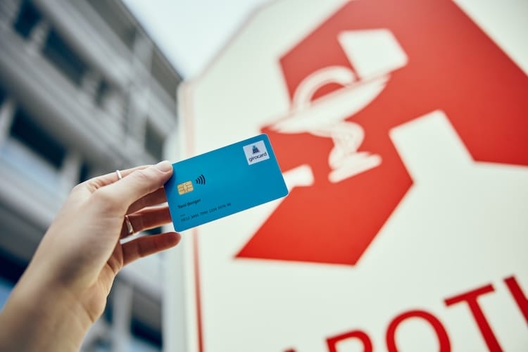 Eine Hand hält eine blaue girocard der Musterbank, im Hintergrund ist ein Apothekenschild zu sehen.
