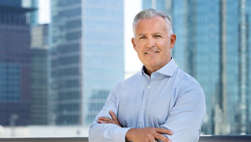 Geschäftsmann mit grauen Haaren, der in einem modernen urbanen Umfeld vor Hochhäusern steht und lächelt, während er die Arme verschränkt.
