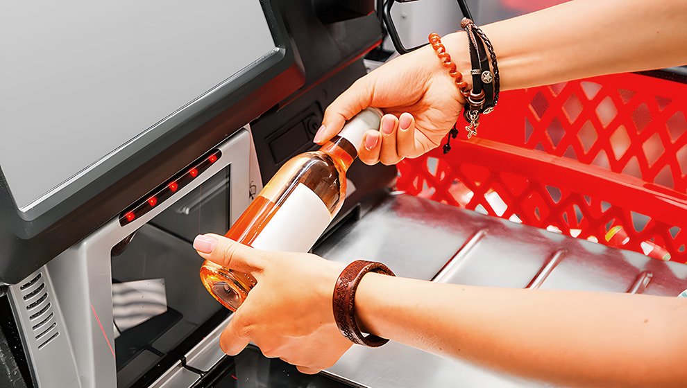 Eine Person scannt eine Flasche an einem Selbstbedienungskassenautomaten im Supermarkt, während der Einkaufswagen im Hintergrund sichtbar ist.