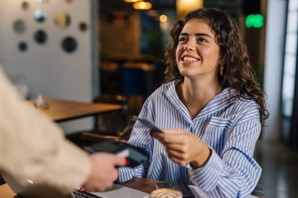 Eine Frau sitzt in einem Café und hält ihre girocard an ein Bezahlterminal und lächelt