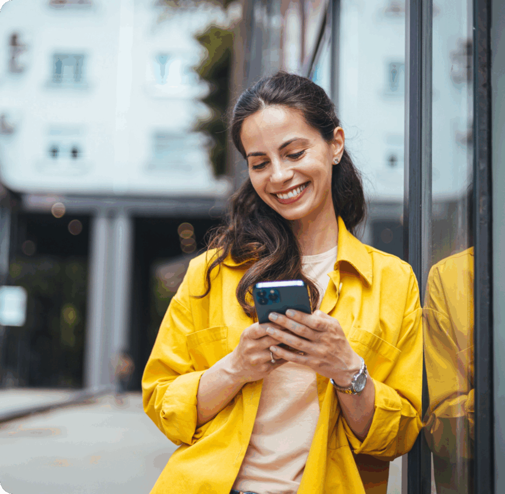Eine junge, braunhaarige Frau in gelber Jacke schaut lächelnd auf das Display ihres Smartphones.