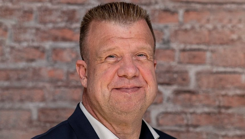 Porträt eines lächelnden Mannes mit kurzen, gestuften Haaren und einem hellen Hemd, vor einer Backsteinwand. Der Mann trägt einen dunklen Blazer und vermittelt einen freundlichen Eindruck.