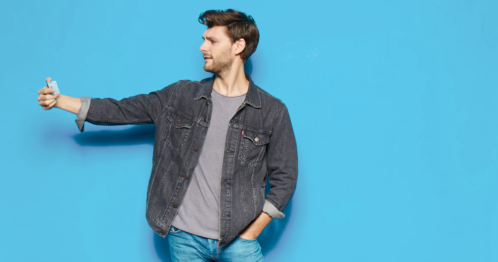 Junger Mann in einer Jeansjacke hält ein kleines Fläschchen in der Hand vor einem hellblauen Hintergrund. Er schaut neugierig auf das Fläschchen und vermittelt einen modernen, lässigen Stil.