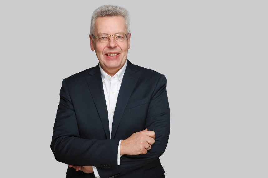 Portrait eines lächelnden Mannes in einem dunklen Anzug mit Brille, der mit verschränkten Armen vor einem grauen Hintergrund steht.